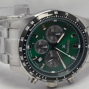 Könnte beinhalten: Edelstahl-Chronographenuhr von Seiko mit grünem Zifferblatt. Die Uhr hat eine schwarze Lünette mit weißen Markierungen, drei Hilfszifferblätter und ein Datumsfenster. Das Armband ist ebenfalls aus Edelstahl.