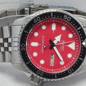 Peut inclure: Une montre automatique Seiko en acier inoxydable avec un cadran rouge et une lunette noire rotative. La montre présente des index, des aiguilles blancs et le jour de la semaine affiché. Le bracelet est de style maillon argenté.