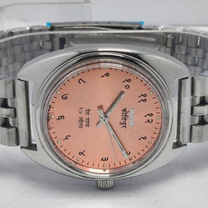 May include: A silver-toned wristwatch with a peach-colored face and a matching silver band. The watch face features Arabic numerals and text in a foreign language. The watch has a classic, elegant design.