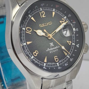 May include: A stainless steel Seiko watch with a black dial. The watch face features gold-colored hands and markers, with the brand name and "Automatic 20BAR" text. The watch has a date display and a stainless steel band.