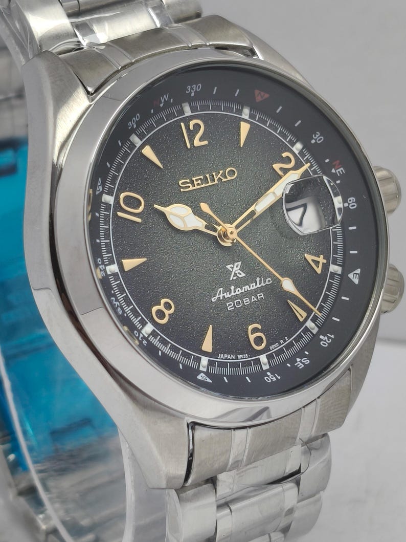 May include: A stainless steel Seiko watch with a black dial. The watch face features gold-colored hands and markers, with the brand name and "Automatic 20BAR" text. The watch has a date display and a stainless steel band.