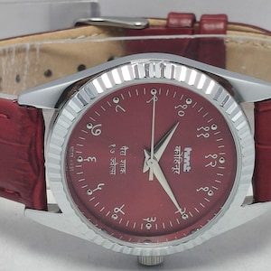 May include: A wristwatch with a burgundy dial and a silver-colored bezel. The watch has a burgundy leather band with a crocodile pattern. The watch face displays Arabic numerals and hands. The watch is a classic accessory.