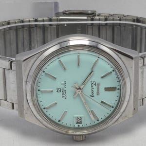 Puede incluir: Un reloj de pulsera plateado con esfera de color verde menta claro, con marcadores de hora y manecillas plateadas. La esfera muestra el texto Tareeq, Para Shock y 17 Jewels, con una ventana de fecha en la posición de las seis y correa metálica.