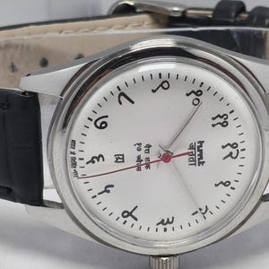 May include: A silver wristwatch with a white face and black Arabic numerals. The watch has a black leather band and a silver case. The watch hands are red. The watch face has text in a foreign language.