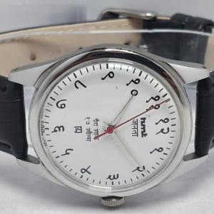 May include: A silver-toned wristwatch with a white face and black Arabic numerals. The watch has a black leather band and a red second hand. The watch face includes text in a foreign language.