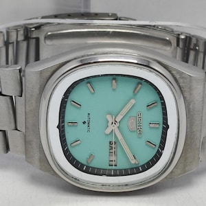 May include: A stainless steel Seiko 5 automatic wristwatch featuring a cushion-shaped case, a light turquoise dial with silver-tone baton indices, luminous hands, and a day-date window at the three o'clock position, paired with a matching link bracelet.