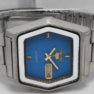 May include: A stainless steel Seiko 5 automatic wristwatch featuring a hexagonal case, a vibrant blue dial with gold-tone hands and markers, and a day-date window displaying SUN 14. The watch includes a matching silver-tone metal link bracelet.
