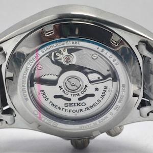 May include: Close-up of the back of a stainless steel Seiko watch. The watch mechanism is visible through a clear glass, showcasing intricate gears and the text "SEIKO TWENTY-FOUR JEWELS JAPAN". The watch is water resistant to 20BAR.