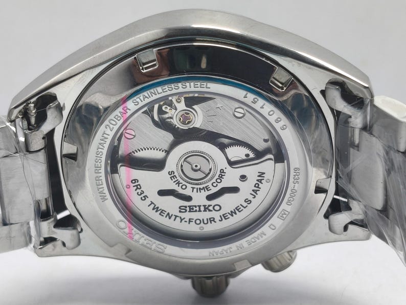 May include: Close-up of the back of a stainless steel Seiko watch. The watch mechanism is visible through a clear glass, showcasing intricate gears and the text "SEIKO TWENTY-FOUR JEWELS JAPAN". The watch is water resistant to 20BAR.