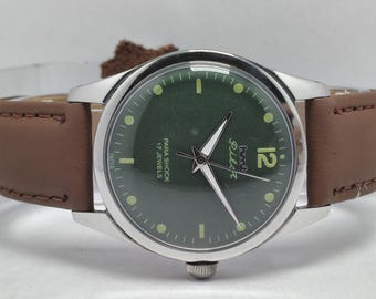 May include: A silver-cased wristwatch with a dark green face and brown leather band. The watch face features white hands, hour markers, and the number 12. The watch is marked "Para Shock 17 Jewels" and "Pilot".