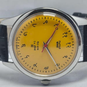 May include: A vintage wristwatch with a silver-tone case and a yellow dial. The watch features black Arabic numerals and red hands. It has a black leather band. The watch face includes text in a foreign language.