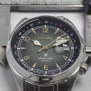 May include: A stainless steel Seiko automatic watch with a dark green dial and gold-tone hands and markers. The watch features a date display at the 3 o'clock position and a rotating bezel with compass markings. The watch is shown with a measuring tool.