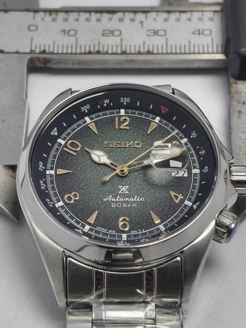May include: A stainless steel Seiko automatic watch with a dark green dial and gold-tone hands and markers. The watch features a date display at the 3 o'clock position and a rotating bezel with compass markings. The watch is shown with a measuring tool.