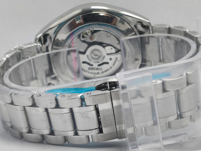 May include: Close-up of a silver-toned wristwatch with a metal band. The watch's back is visible, showcasing the inner workings and the text "SEIKO TWENTY-FOUR JEWELS JAPAN". The watch is secured with a clear plastic band.