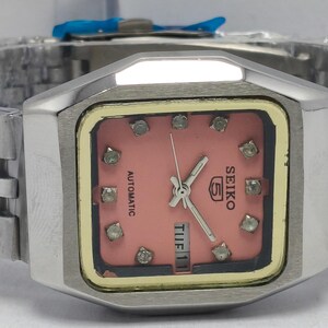 May include: A silver Seiko 5 automatic watch with a pink square face. The watch has a silver strap, a gold-coloured inner frame, and diamond-shaped hour markers. The watch face displays the day and date.