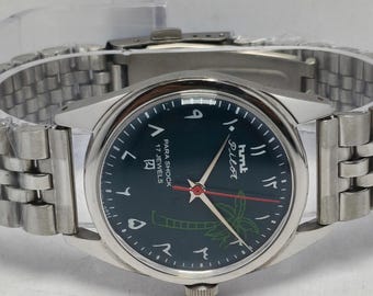 May include: A silver-toned wristwatch with a dark teal face and a matching silver band. The watch face features Arabic numerals, a palm tree graphic, and the word "Pilot". The watch has a second hand with a red tip.