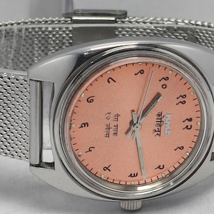 May include: A silver-toned wristwatch with a mesh band and a salmon-colored face. The watch face features black numerals and hands, with text in a foreign language. The watch is a classic style, suitable for everyday wear.