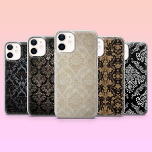 Può includere: Cinque custodie per telefono con motivi ornati in stile damascato. Le custodie presentano una varietà di colori, tra cui nero, oro e crema, con un bordo trasparente. Le custodie sono disposte in fila su uno sfondo rosa.