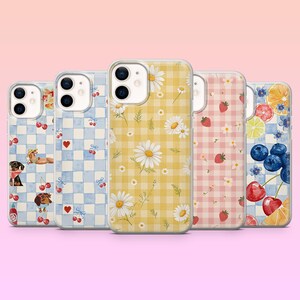 May include: A selection of phone cases with diverse designs. The cases display patterns such as gingham, daisies, and fruit. Some cases have illustrations of dogs, cherries, and strawberries. The cases are arranged in a row against a pink backdrop.