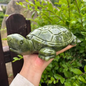 May include: A detailed, green stone turtle sculpture. The turtle is carved with a textured shell and visible legs and head. The sculpture is held in a hand, with a background of green foliage.