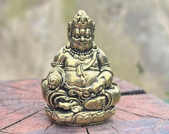 May include: A small, detailed brass statue of a seated deity. The figure has a serene expression, ornate headdress, and is adorned with necklaces and a draped garment. The statue is on a wooden surface, with a blurred green background.