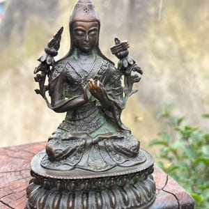 May include: Bronze statue of a seated figure with hands in prayer. The figure wears a detailed headdress and ornate clothing. Two floral elements flank the figure. The statue is on a decorative base, showcasing intricate patterns and textures.