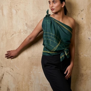 Puede incluir: Un top verde oscuro de un solo hombro con un patrón de cuadrícula amarilla y un detalle de nudo atado. El top se combina con pantalones negros. El atuendo se encuentra contra una pared de tonos neutros.
