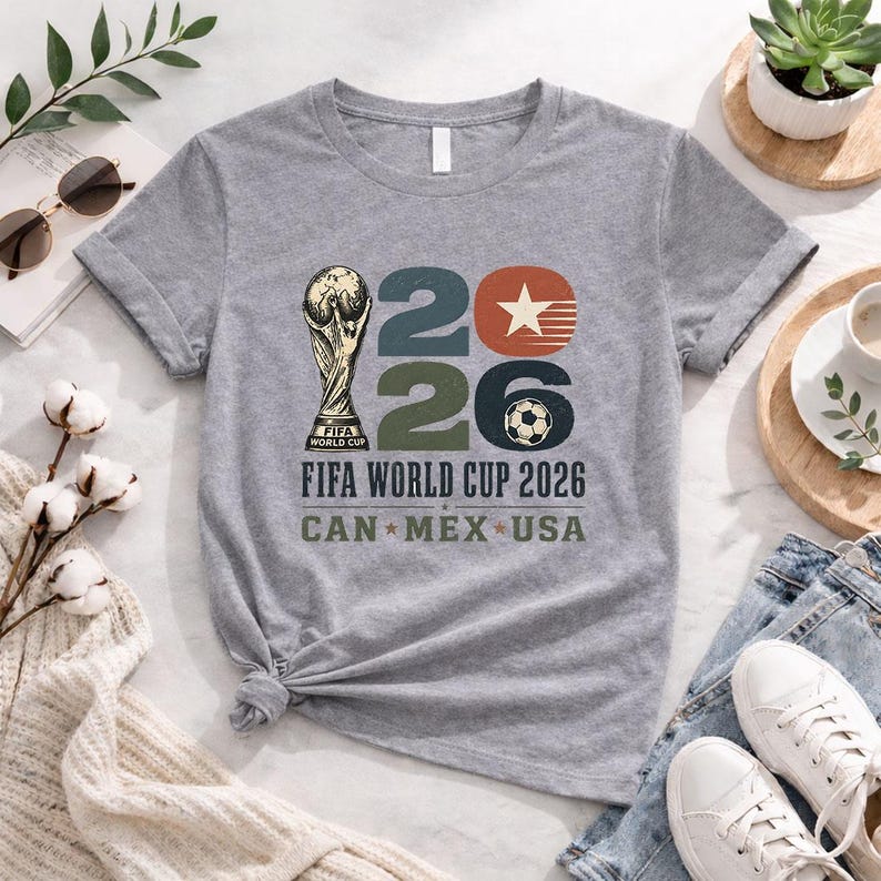 May include: A gray t-shirt featuring the text "2026 FIFA WORLD CUP" with a trophy graphic and the text "CAN * MEX * USA". The shirt has short sleeves and a crew neck.