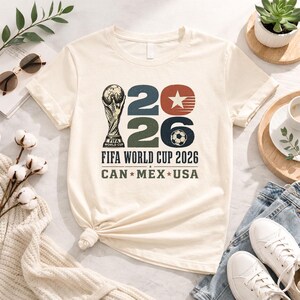May include: Cream-colored t-shirt featuring a graphic for the FIFA World Cup 2026. The design includes the FIFA trophy, the numbers "2026", a star, and a soccer ball. The text "CAN * MEX * USA" is also printed on the shirt.