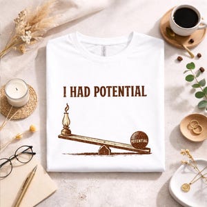 Puede incluir: Camiseta blanca con el texto marrón "I HAD POTENTIAL". Una ilustración marrón muestra un balancín con una lámpara y una bola con la etiqueta "POTENTIAL". La camiseta está plana.