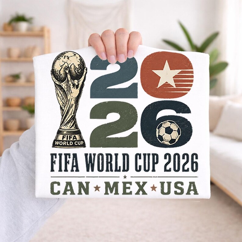 May include: White t-shirt featuring the FIFA World Cup trophy and the text "2026 FIFA WORLD CUP CAN * MEX * USA". The design includes the numbers "2026" in blue, green, and black, with a soccer ball graphic.
