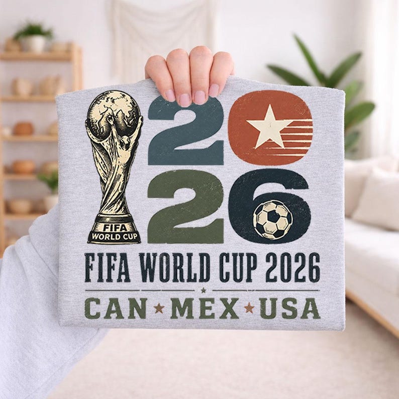 May include: A light gray t-shirt featuring the FIFA World Cup trophy and the text "2026 FIFA WORLD CUP CAN * MEX * USA". The numbers "2026" are in green, brown, and black. A soccer ball graphic is on the number 6.