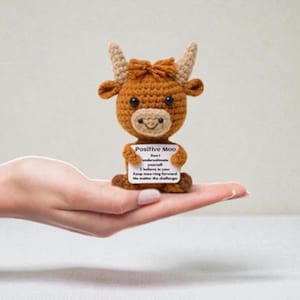 May include: A crocheted brown bull figurine with a sign that reads "Positive Moo." The bull has tan horns and a light brown face. The sign has an inspirational message. The figurine is held in a person's hand.