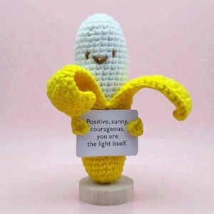 May include: A crocheted banana figurine with a smiling face and yellow peel. The banana holds a small sign that reads "Positive, sunny, courageous, you are the light itself." The figurine is on a wooden base.