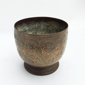 Puede incluir: Un cuenco antiguo de color bronce con intrincados patrones florales y foliares grabados en su superficie. El cuenco tiene un cuerpo redondeado, un borde acampanado y una pequeña base circular. El interior muestra una pátina verde.