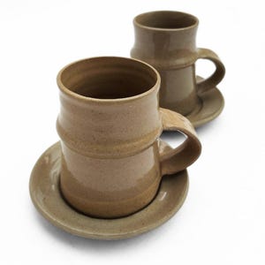 May include: Two handmade ceramic mugs with matching saucers. The mugs are a light brown color with a textured surface and a cylindrical shape. Each mug has a handle and sits on a round saucer. The background is white.