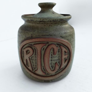 May include: A ceramic rice jar with a lid. The jar is a muted green color with a reddish-brown oval design on the front that reads "RICE". The jar has a textured surface and a small handle on the lid.
