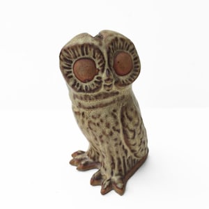 May include: A ceramic owl figurine featuring a mottled olive-green and brown glaze. The owl has large, circular, smooth brown eyes and textured, carved feather details across its body, with a small beak and three-toed feet visible at the base.