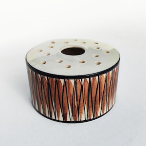 Puede incluir: Un ranero de cerámica con un diseño circular. La parte superior es de color gris claro con múltiples agujeros para los tallos de las flores. Los lados están decorados con un patrón geométrico marrón y negro. La base es negra.