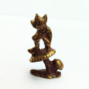 Könnte beinhalten: Kleine, messingfarbene Figur einer Kreatur mit spitzen Ohren, die sitzt. Die Figur sitzt auf einem Pilz, dessen Stiel und Basis ebenfalls sichtbar sind. Das Design deutet auf ein skurriles oder Fantasy-Thema hin.