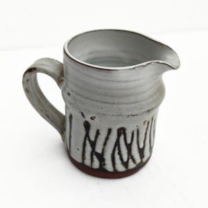 Op de afbeelding: Handgemaakte keramische kan met een grijze glazuur en een donkerbruine basis. De kan heeft een handvat en een tuit om uit te schenken. De buitenkant heeft een donker, abstract patroon. Deze rustieke kan is perfect voor het serveren van dranken.