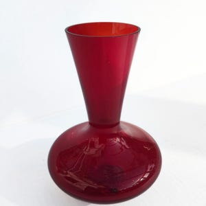 Puede incluir: Un jarrón de cristal rojo rubí con base redondeada y cuello cónico acampanado. El jarrón tiene un acabado liso y brillante y está sobre un fondo blanco. El diseño es sencillo y elegante.