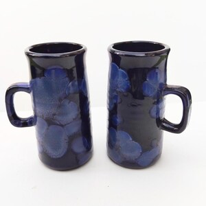 Può includere: Due tazze alte in ceramica con una smaltatura di base blu scuro che presenta motivi cristallini in tonalità di blu più chiare. Ogni tazza ha un corpo cilindrico e un manico laterale, con una finitura lucida e formazioni cristalline floreali.