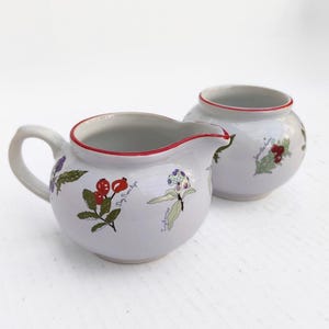 May include: A white ceramic creamer and sugar bowl set. The creamer has a handle and spout, and the sugar bowl is round. Both pieces have a red rim and are decorated with botanical illustrations of berries and flowers.