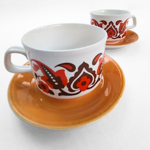 Może przedstawiać: Dwie białe ceramiczne filiżanki do herbaty z pasującymi pomarańczowymi spodkami. Filiżanki mają dekoracyjny wzór w kolorze czerwonym, brązowym i czarnym. Filiżanki są na białym tle.