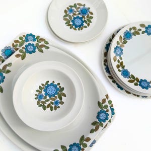 Puede incluir: Un conjunto de vajilla de cerámica blanca con un diseño floral azul. El juego incluye platos, cuencos y fuentes. El patrón floral presenta flores azules con hojas verdes y marrones. Los platos son redondos y cuadrados, y los cuencos son redondos.