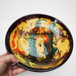 May include: A decorative ceramic bowl with a vibrant, abstract design. The bowl features a dark brown rim and a swirling pattern of yellow, red, green, and brown colors. The bowl is held by a hand.