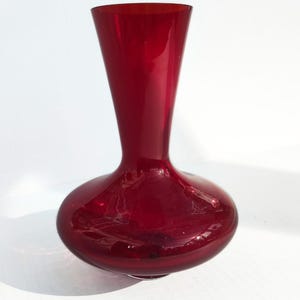 May include: A deep red glass vase with a rounded base and a flared top. The vase has a smooth, glossy finish. The design is simple and elegant. The vase casts a shadow on the white surface below.