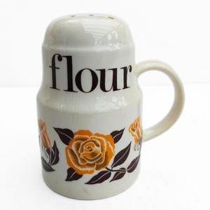 Könnte beinhalten: Ein weißer Keramik-Mehlstreuer mit Griff und Löchern oben. Das Wort "flour" ist schwarz aufgedruckt. Verziert mit orangefarbenen Rosen und braunen Blättern. Ein Küchenartikel.