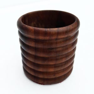 Può includere: Una tazza o contenitore in legno marrone scuro con un design a coste e impilato. La tazza ha un bordo liscio e arrotondato e una forma cilindrica. La venatura del legno è visibile, aggiungendo consistenza al pezzo. Il contenitore poggia su una superficie bianca.